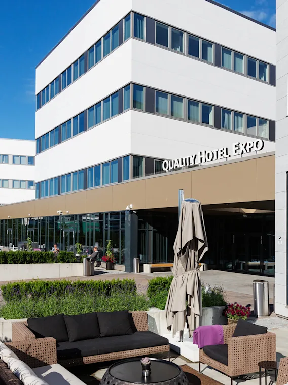 Quality Hotel Expo-bygning med moderne eksteriør, med glass og betong. Utendørs sitteområde med rottingmøbler og parasoller. Omgivende grøntområder og klar blå himmel.