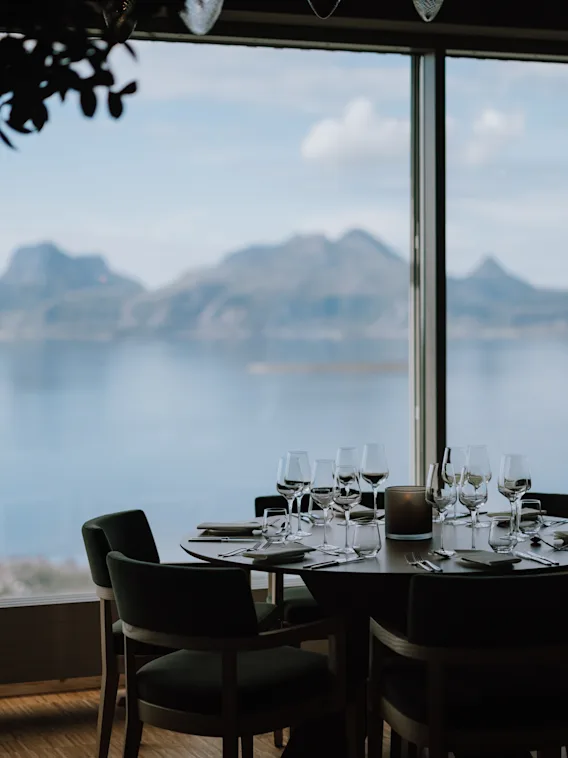 Ett bord med snyggt arrangerade glas och bestick har panoramautsikt över berg och vatten genom ett stort fönster. En person står i närheten i en restaurangmiljö.