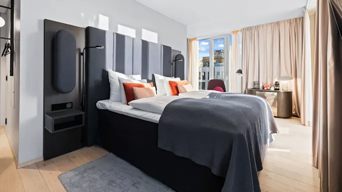 A large bed with gray and orange pillows stands in a modern, sunlit room. Floor-to-ceiling windows and light curtains create an airy, inviting atmosphere.