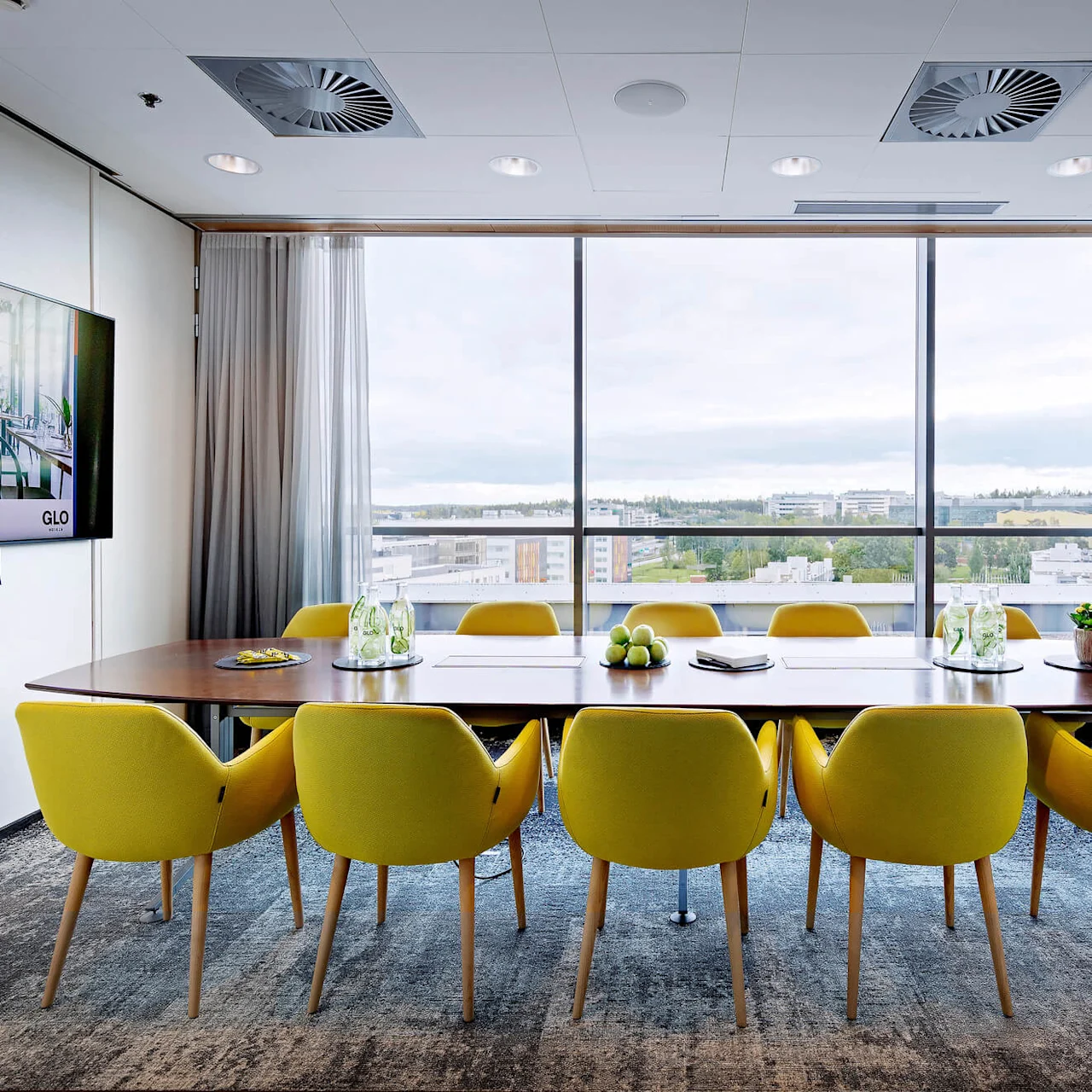 A conference room features a long table with yellow chairs and water bottles, overlooking a cityscape through large windows. A wall-mounted screen displays "GLO."