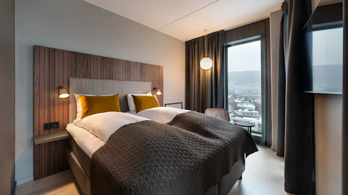 A modern hotel room features a double bed with mustard pillows and a brown quilt, framed by wooden paneling. A window offers views of snowy rooftops in an urban setting.
