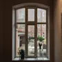 Et vindue vender ud mod en gårdhave med udendørs siddepladser og murstensbygninger. En plante og vase står på vindueskarmen, med kunstværk i nærheden.