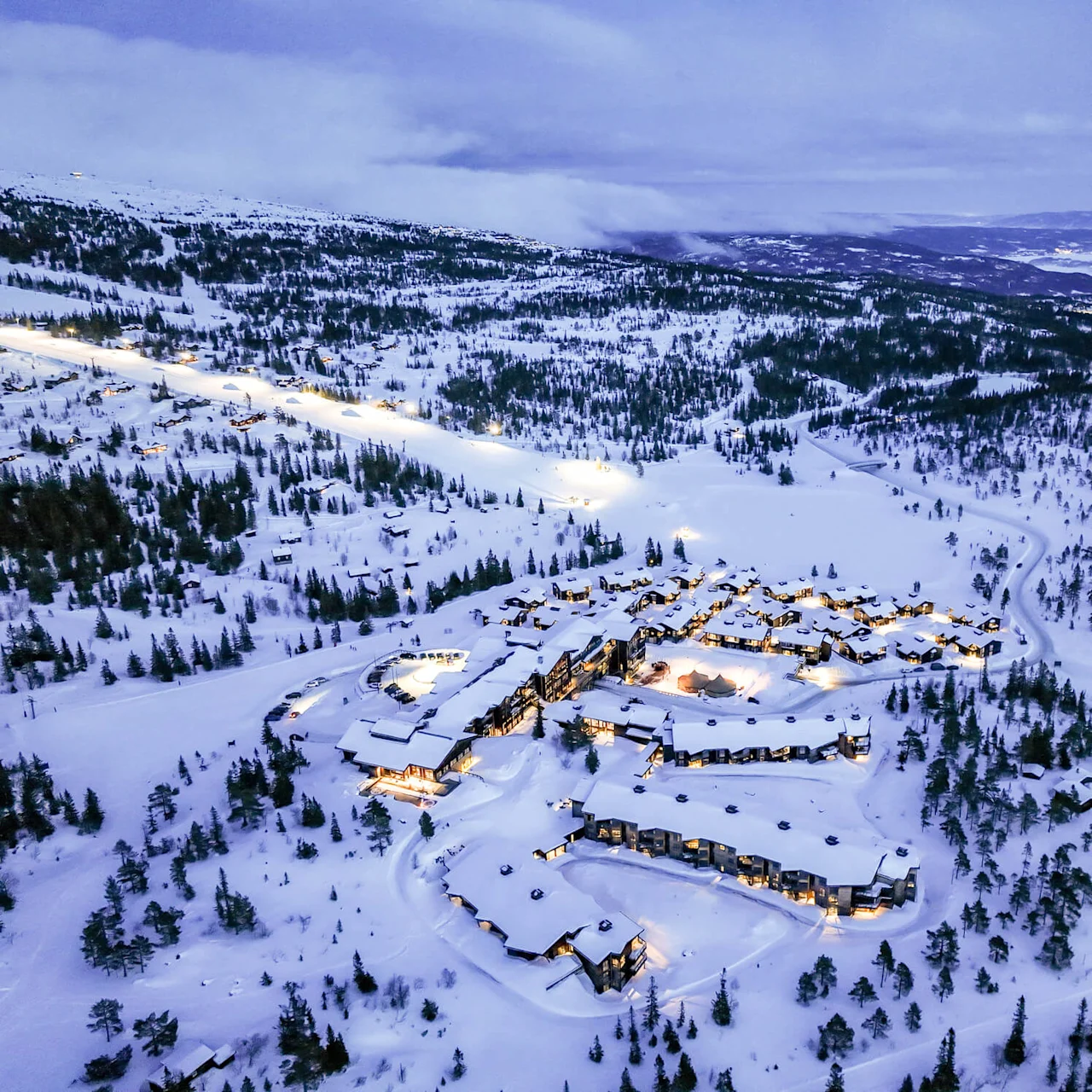 Snow-covered buildings emit warm light, nestled in a vast, snowy landscape surrounded by trees and mountains. Twisting roads connect the structures, under a subdued, cloudy sky. 