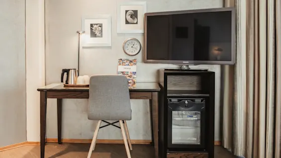 Ett hotellrum med ett skrivbord, stol och en dryckesstation, tillsammans med en TV och minibar. En broschyr för TROMSO ligger på skrivbordet.