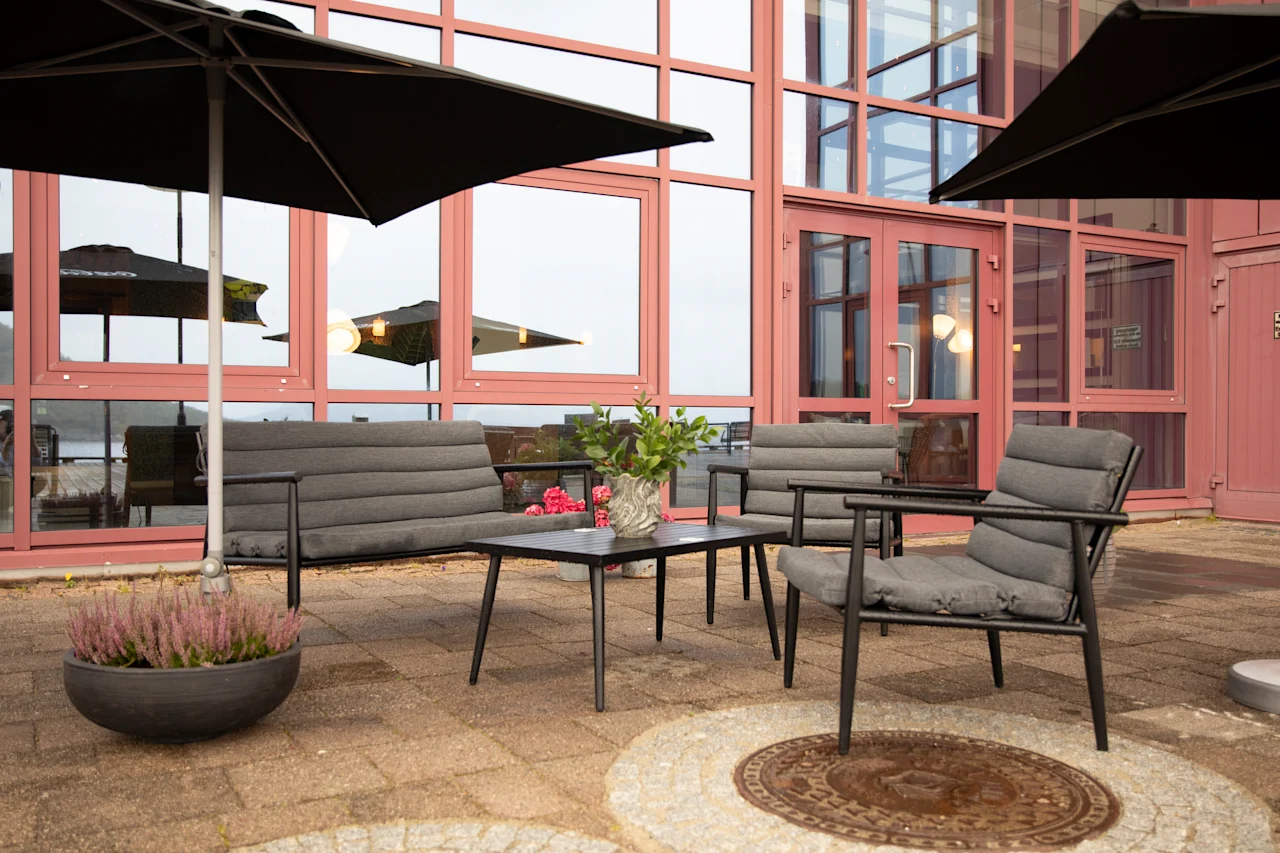 Kutsuva ulkoterassi mukavilla harmailla istuimilla, pöydällä ja aurinkovarjoilla, modernin rakennuksen edessä suurilla ikkunoilla.