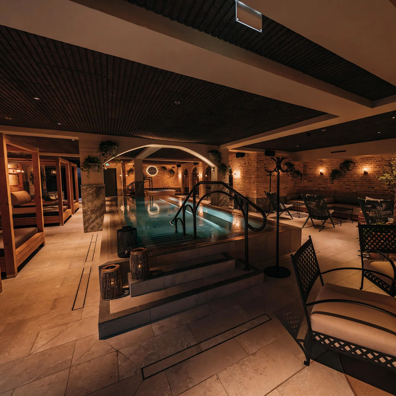 A swimming pool surrounded by lounge chairs and dim lighting in a cozy, indoor spa setting with brick walls and plants.