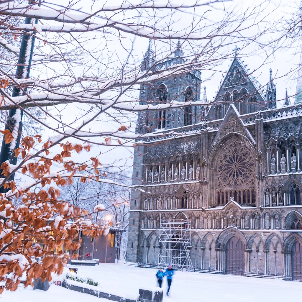 En storslået, snedækket katedral med indviklede detaljer, indrammet af nøgne træer med et par tilbageværende brune blade. To personer går i sneen.
