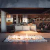 En moderne lounge har en rektangulær ildsted i midten med flammer over sten, omgitt av stoler og sofaer. Miljøet er en stilren innendørs arena med varm belysning.