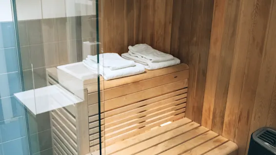 A wooden sauna bench is neatly stacked with white towels, situated in a small, glass-enclosed sauna room, surrounded by light wooden paneling and adjacent tiled walls.