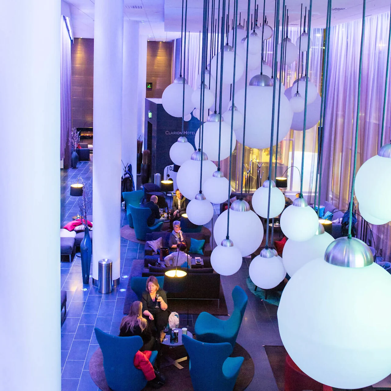 Stora sfäriska lampor hänger från ett högt tak och lyser upp en modern lounge där människor sitter i blå stolar och pratar. Gardiner och höga vita pelare förstärker den stiliga atmosfären. "Clarion Hotel" text syns.