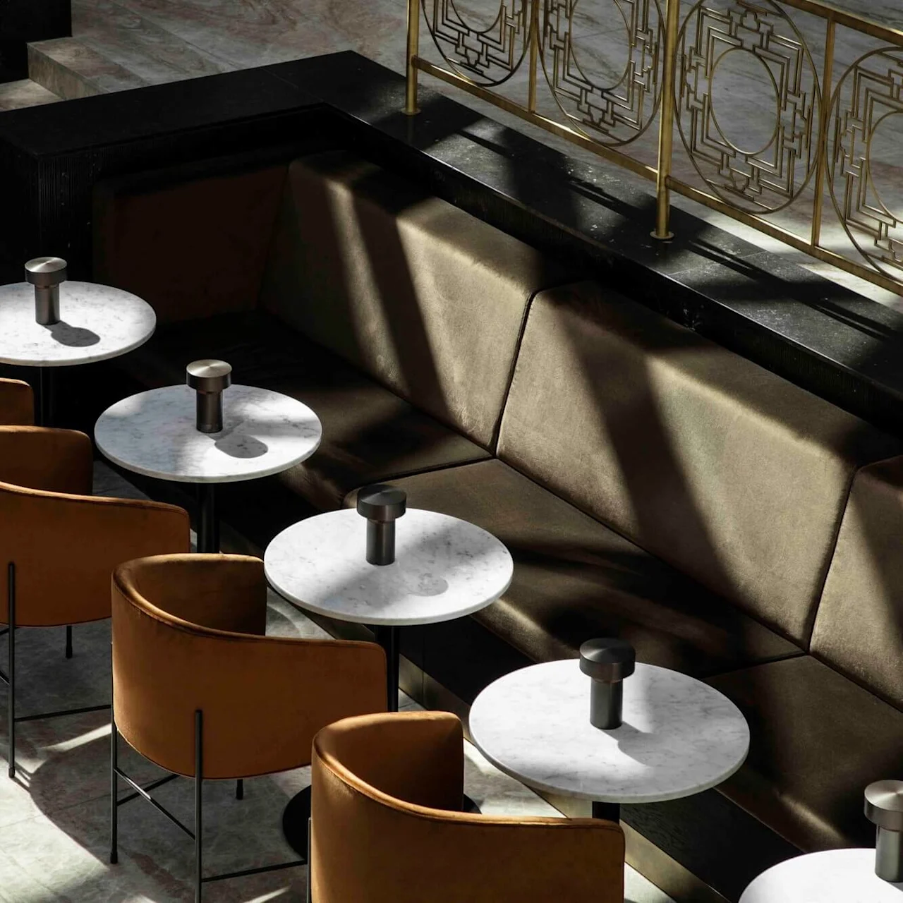 Tables with round marble tops are placed in a row, accompanied by brown upholstered chairs. The setting is a stylish lounge area with soft lighting.