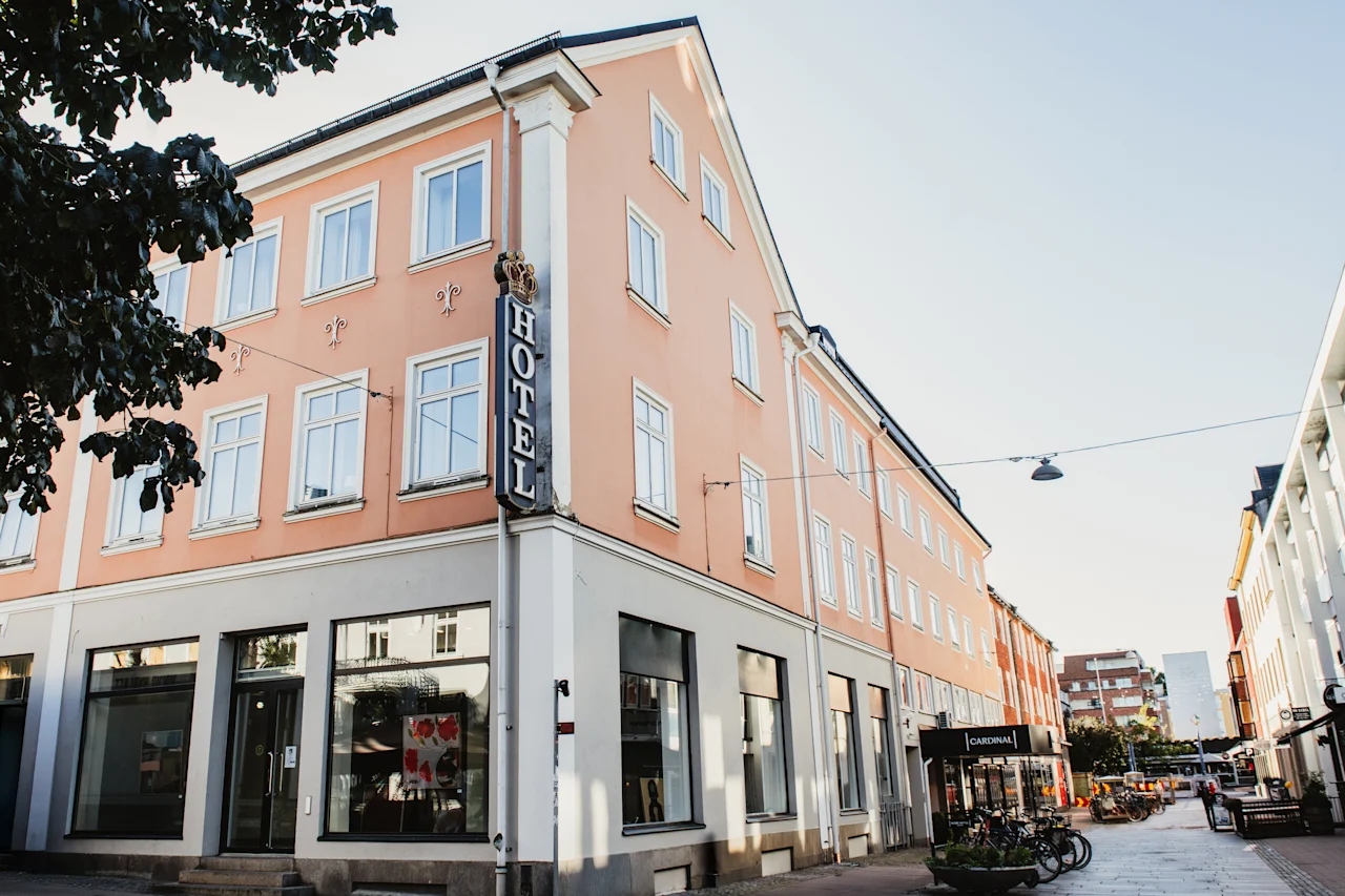 Persikanvärinen hotellirakennus, jonka kulmassa on näkyvä pystysuora HOTEL-kyltti, sijaitsee aurinkoisella kadulla muiden rakennusten ja pysäköityjen polkupyörien kanssa.