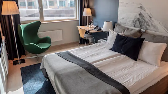 Säng med grå och vita sängkläder i ett modernt hotellrum. En grön fåtölj och lampa står vid fönstret. Ett skrivbord med telefon lutar mot en mörk vägg med ett stort naturfoto.