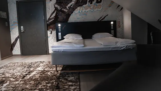 A bed with white bedding sits under an attic ceiling, flanked by lamps. A mural of a dynamic dark figure and Japanese text, "れんゆくもの", decorates the wall.