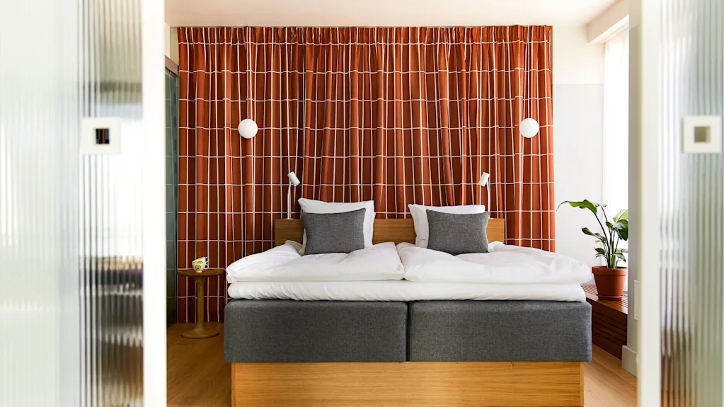 Bed with gray headboard sits against red-patterned curtain. Two white pillows and two gray cushions rest on the bed. A potted plant stands to the right.