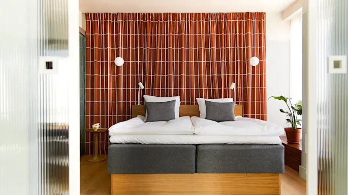 Seng med grå hodegjerde står mot rødt-mønstret gardin. To hvite puter og to grå puter ligger på sengen. En potteplante står til høyre.