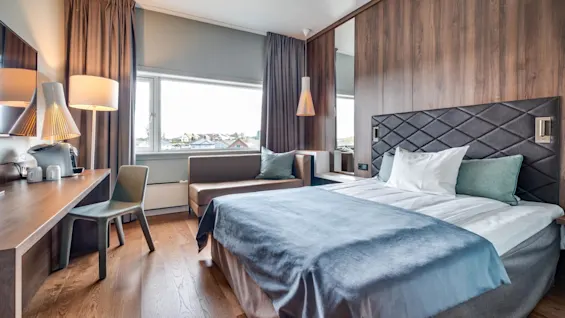 A modern, well-lit hotel room with a large bed, desk, lamps, sitting area, and a window with a view of houses.