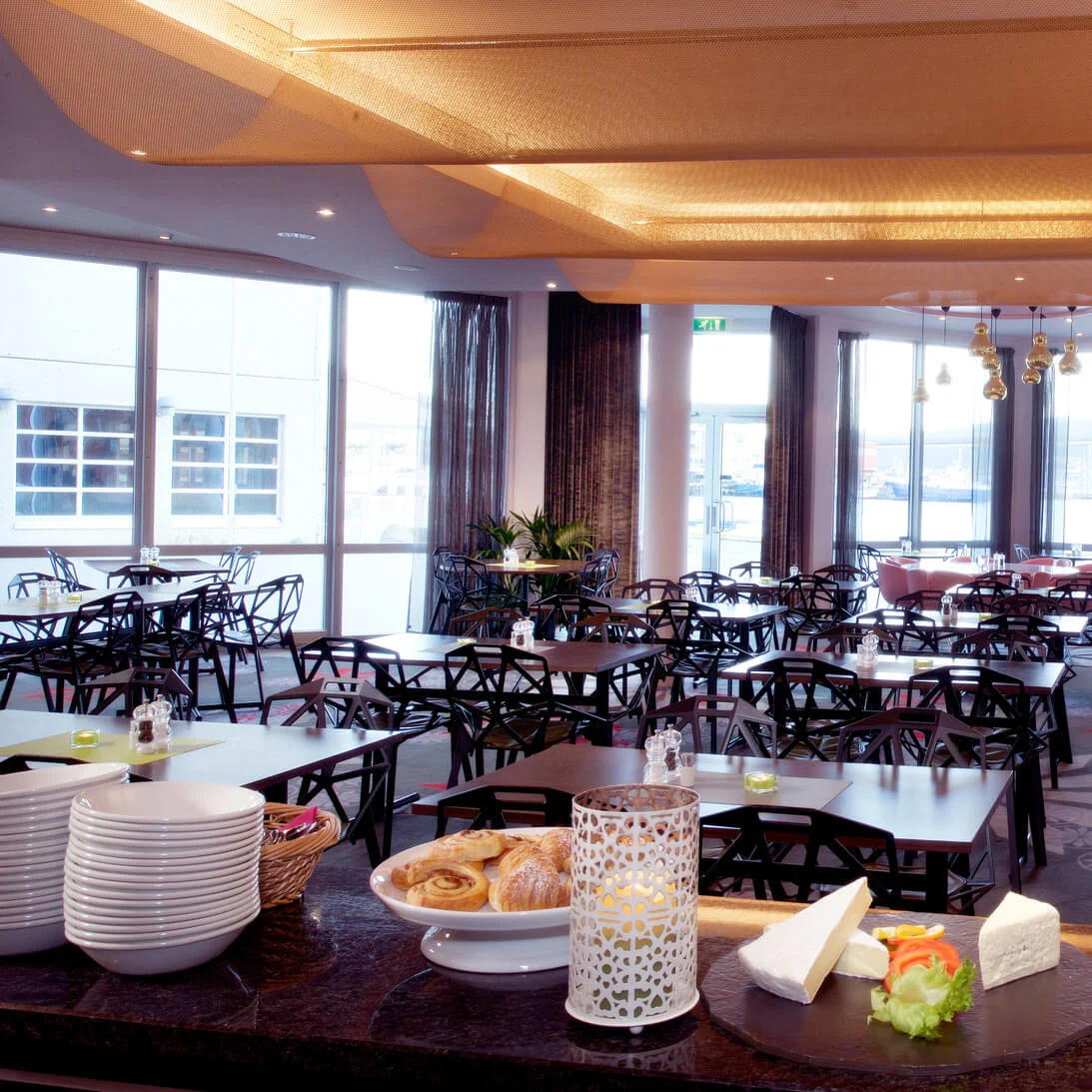 Modern restaurant interior with empty black chairs and tables, ready for service. Large windows provide natural light.