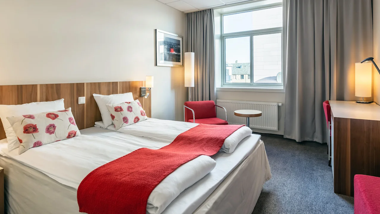 En pent hotelseng med røde detaljer står i et rom med et vindu, grå gardiner, en rød stol og et trebord under varm belysning.
