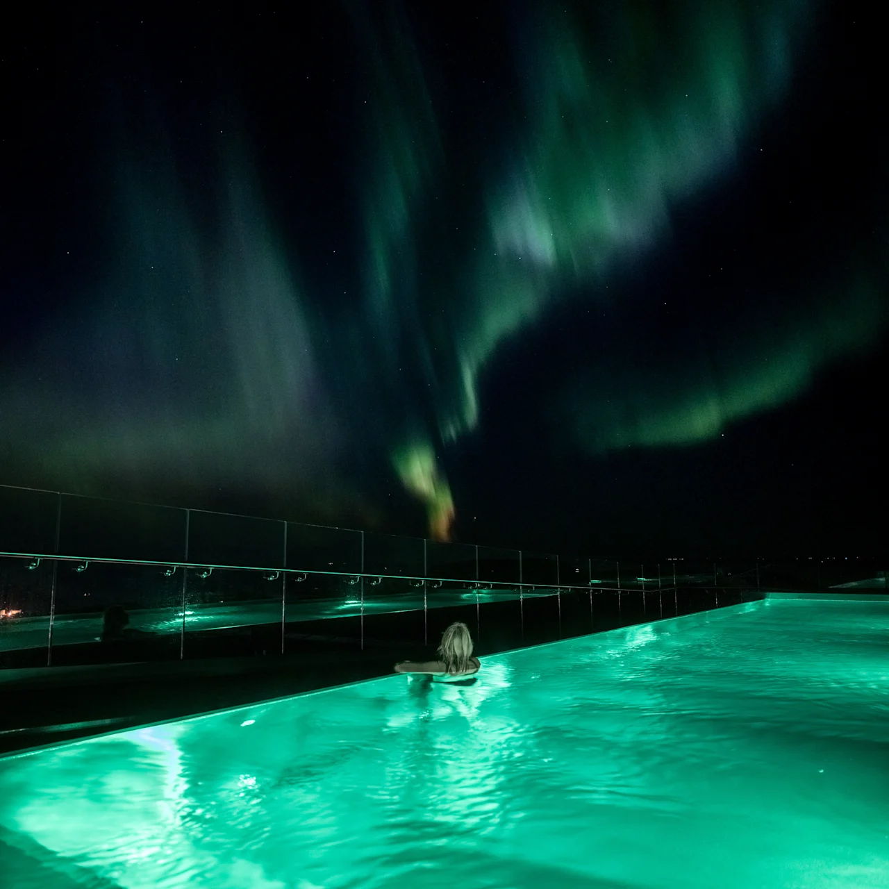 En person nyter et bad i et glødende grønt basseng om natten, med det livlige nordlyset dansende over den mørke himmelen.