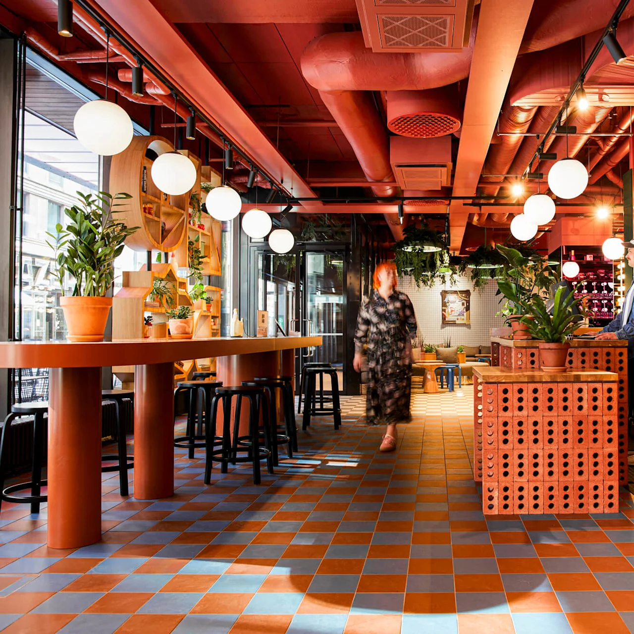 Et moderne café med en person, der går, og en anden ved en disk. Jordnære planter dekorerer træborde blandt stærkt lys og rød-murstensmønstrede vægge.