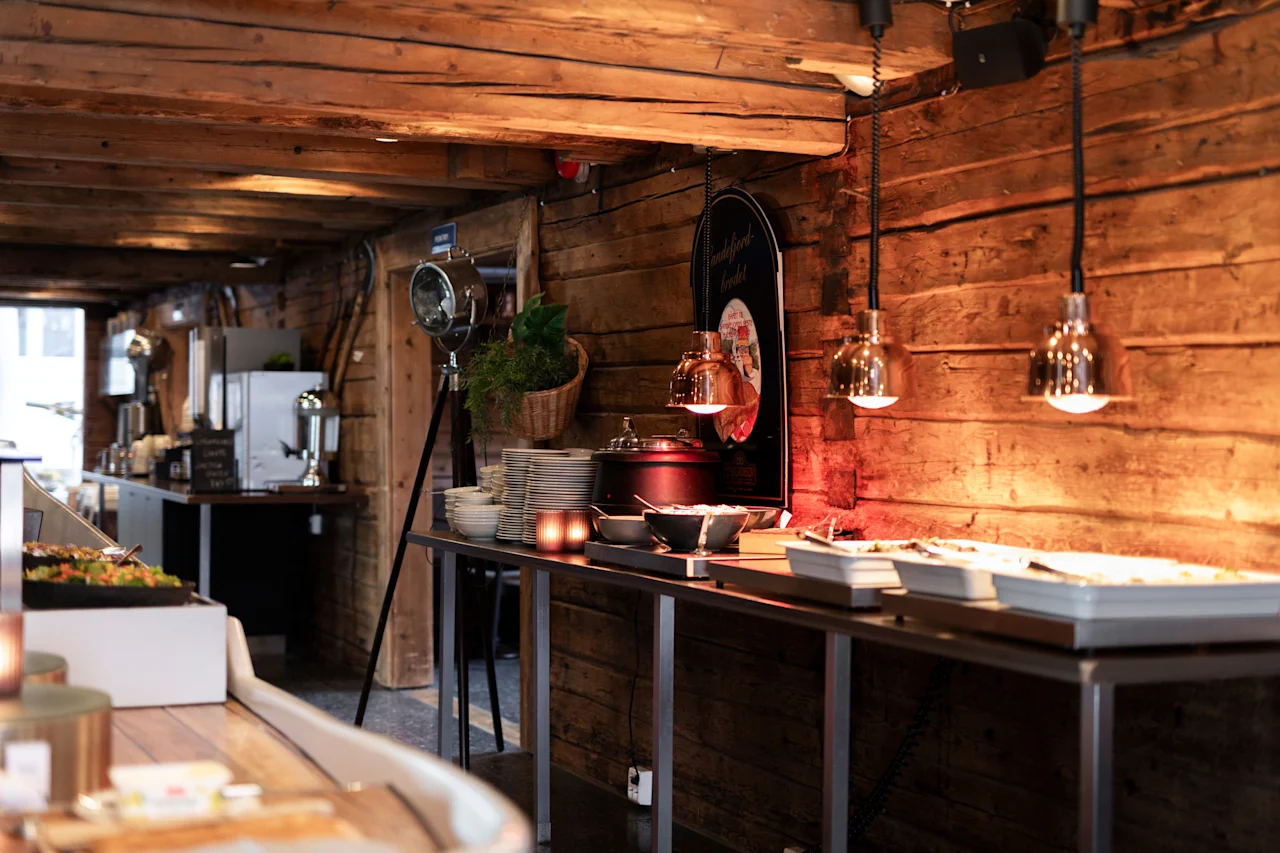Et hyggeligt, rustikt buffetområde med varm belysning, træbjælker og et udvalg af mad lagt ud til gæster.