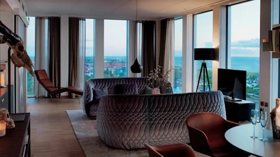 Lyxig grå soffa står i modernt vardagsrum, vänd mot en platt-TV. Stora fönster visar stad och havsutsikt, med elegant inredning inklusive golvlampa och runt bord.