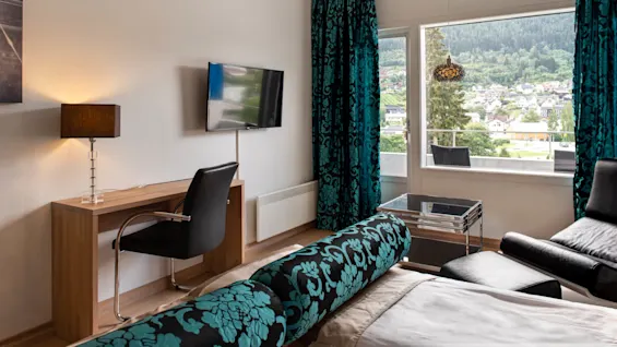 Ett modernt hotellrum med ett skrivbord med en lampa, en väggmonterad TV och ett stort fönster med utsikt över en stad och skogsklädda kullar. Blå mönstrade gardiner lägger till färg.