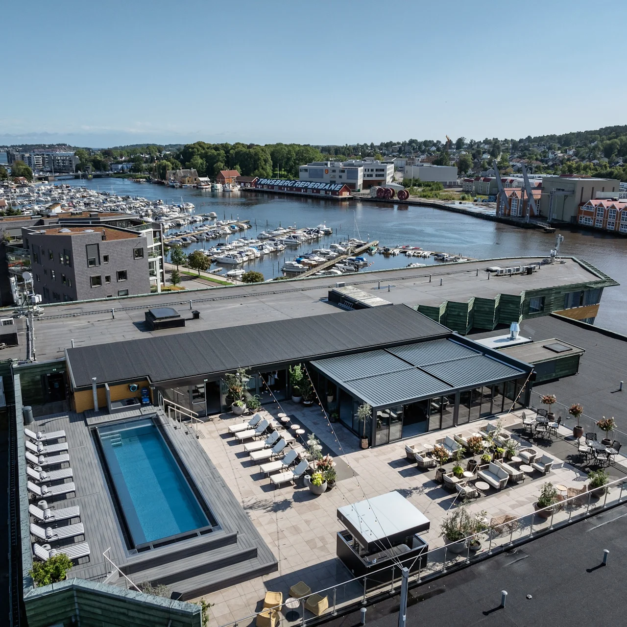En takterrass har en swimmingpool omgiven av solstolar och sittplatser. Den har utsikt över en marina med många båtar vid en flod. Stor skylt lyder "TØNSBERGRØRA PRAMA".