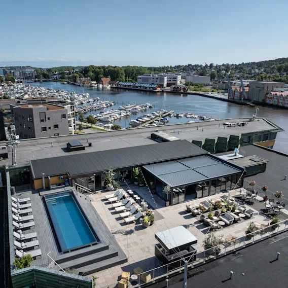 En takterrasse har et svømmebasseng omgitt av solsenger og sitteområder. Den har utsikt over en marina med mange båter langs en elv. Stor skilt leser "TØNSBERGRØRA PRAMA".