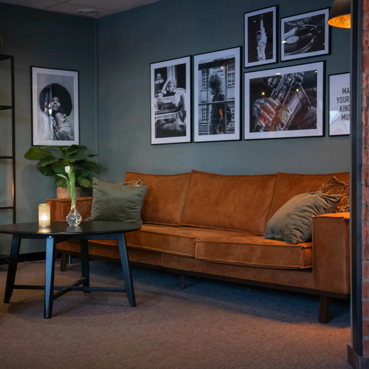 Brown leather sofa in a stylish room, adorned with framed black-and-white photos on the wall. Nearby, a black coffee table holds a vase with a tulip. Ambient lighting provides a cozy atmosphere. Text: "MAKE YOUR KIND OF MUSIC."