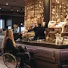 En kvinne i rullestol mottar et kort fra en kasserer i en moderne kafé med dekorativ belysning, en disk med snacks og en naturskjønn utsikt.