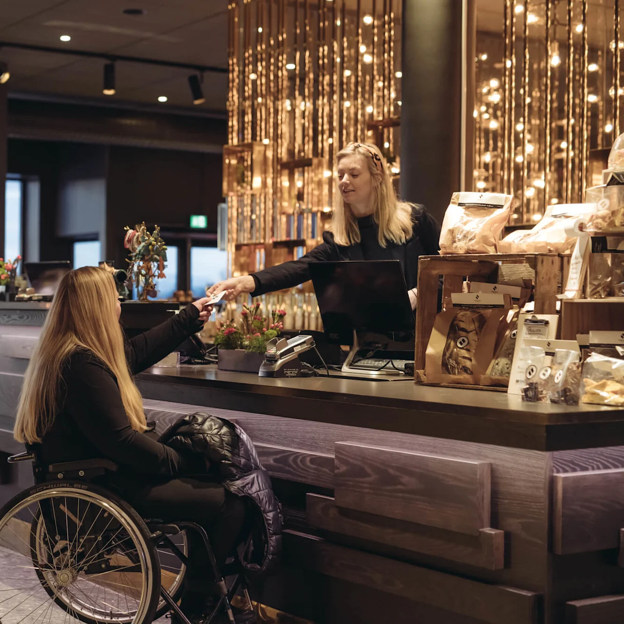 En kvinna i rullstol tar emot ett kort från en kassör i ett modernt café med dekorativ belysning, en disk med snacks och en naturskön utsikt.