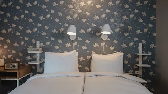 A neatly made twin bed is against a floral-patterned wallpaper. Two wall-mounted lamps hang above. Shelves beside the bed hold books and a vintage radio.