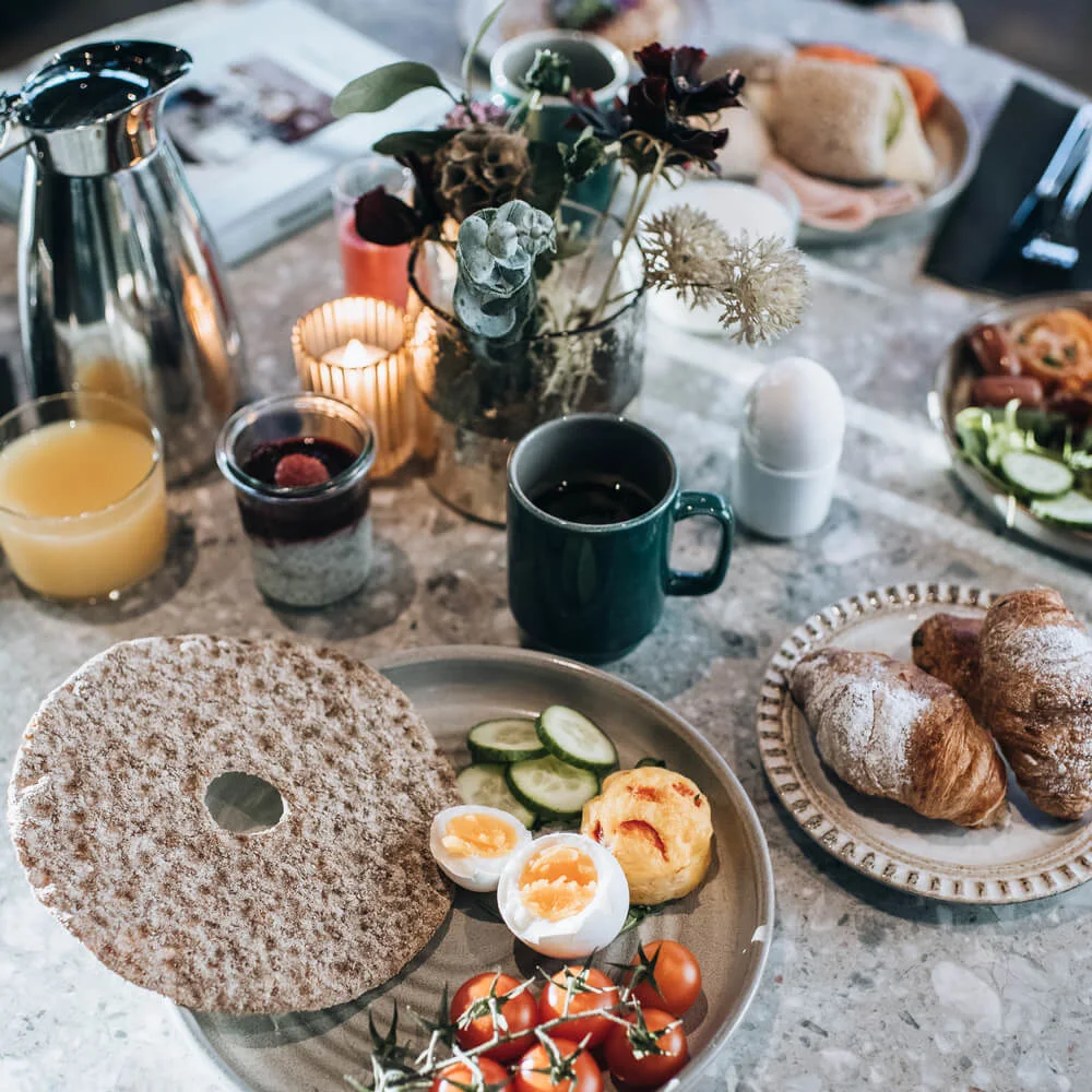 A breakfast spread on a round table features crispbread, soft-boiled eggs, cherry tomatoes, and cucumbers, surrounded by croissants, juice, coffee, flowers, lit candle, and a shiny carafe.