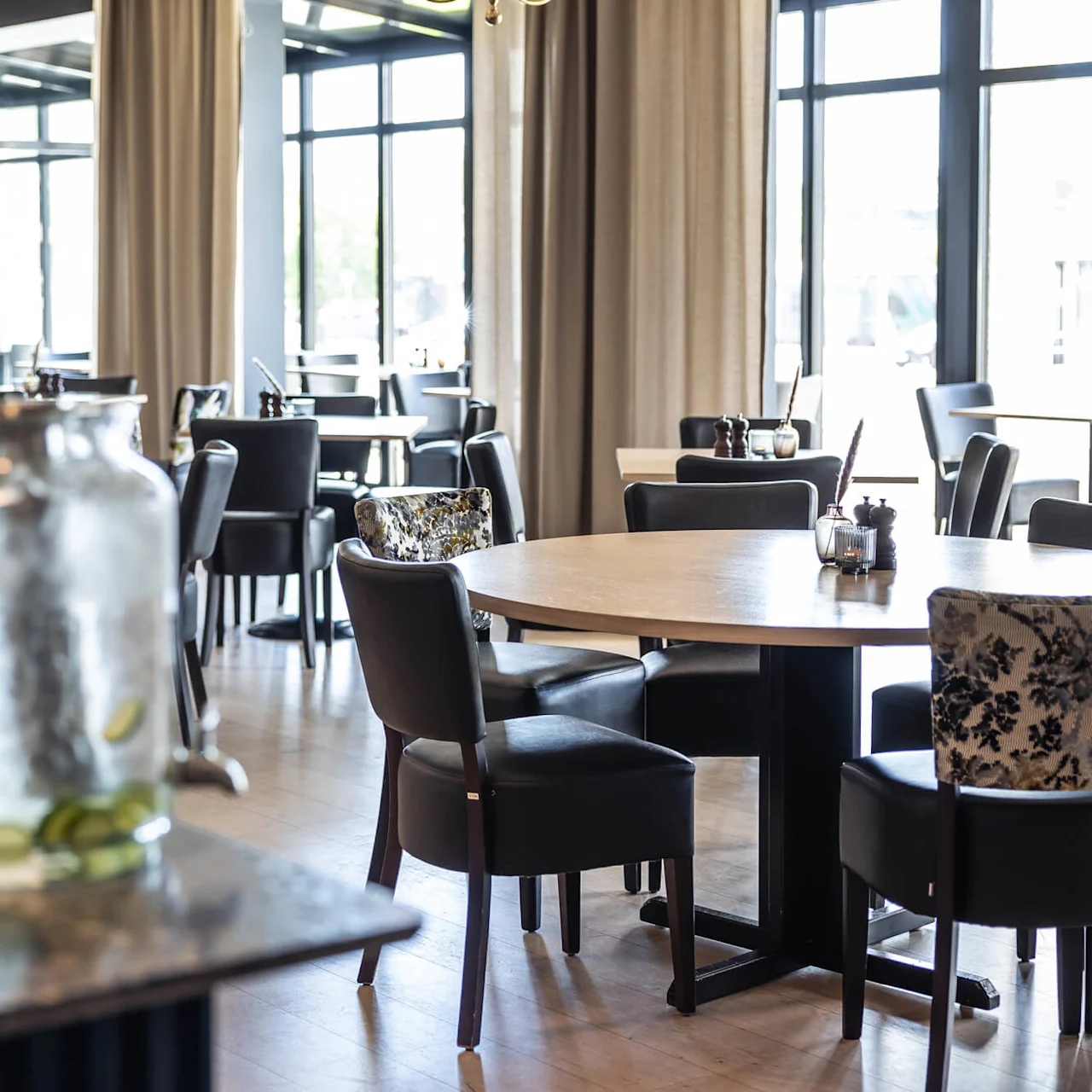 Restaurantborde og stole er arrangeret i et rummeligt spiseområde med store vinduer og elegant indretning, herunder et dekorativt spejl og lysekroner. En skål med frugt og vandkrukke pryder forgrunden.