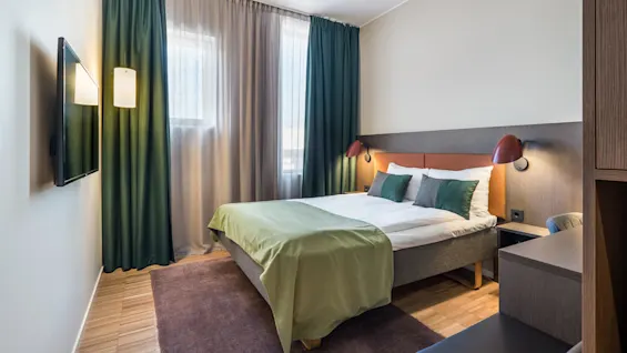 A bed covered with green bedding is in a neatly arranged bedroom. A TV is mounted on the wall beside dark curtains. Soft lighting adds warmth to the space.