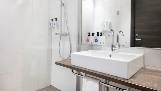 A modern bathroom features a white sink on a wooden counter with a chrome faucet, next to a glass-enclosed shower. Walls are white tile, with soap dispensers and a mirror.