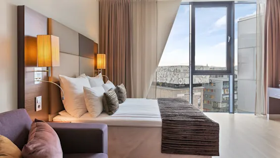 A neatly made bed with decorative pillows is flanked by wall-mounted lamps in a modern hotel room, overlooking city buildings through a large window.