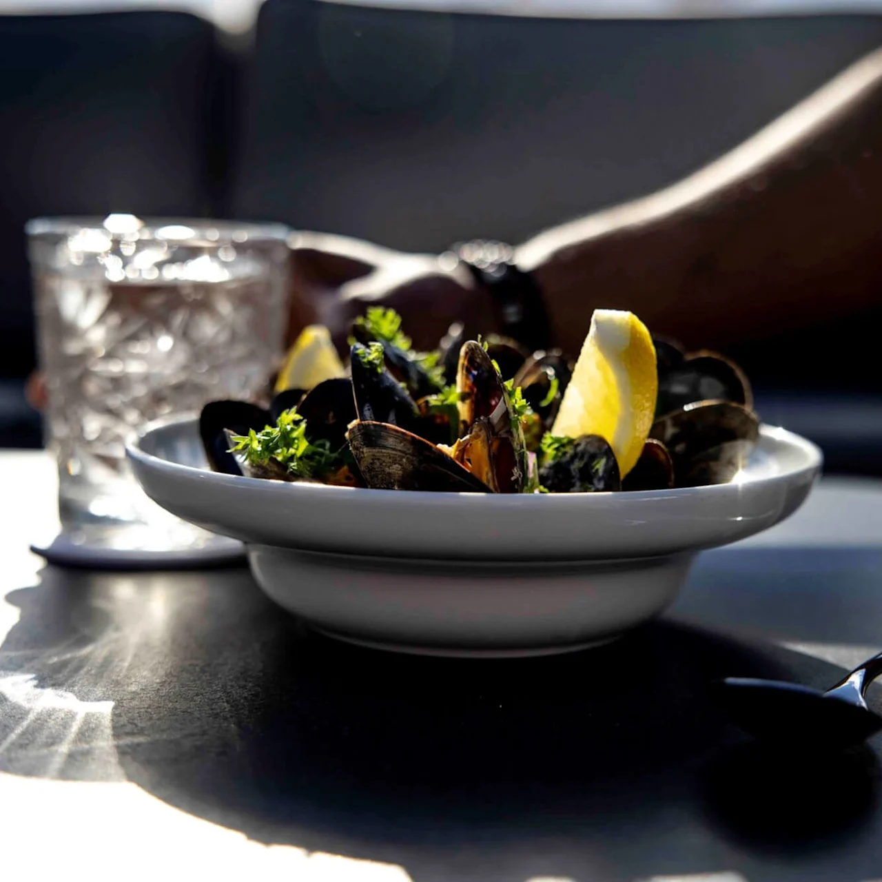 A bowl of mussels garnished with lemon slices and herbs on a table beside a glass of water, with a hand reaching out and a spoon nearby.