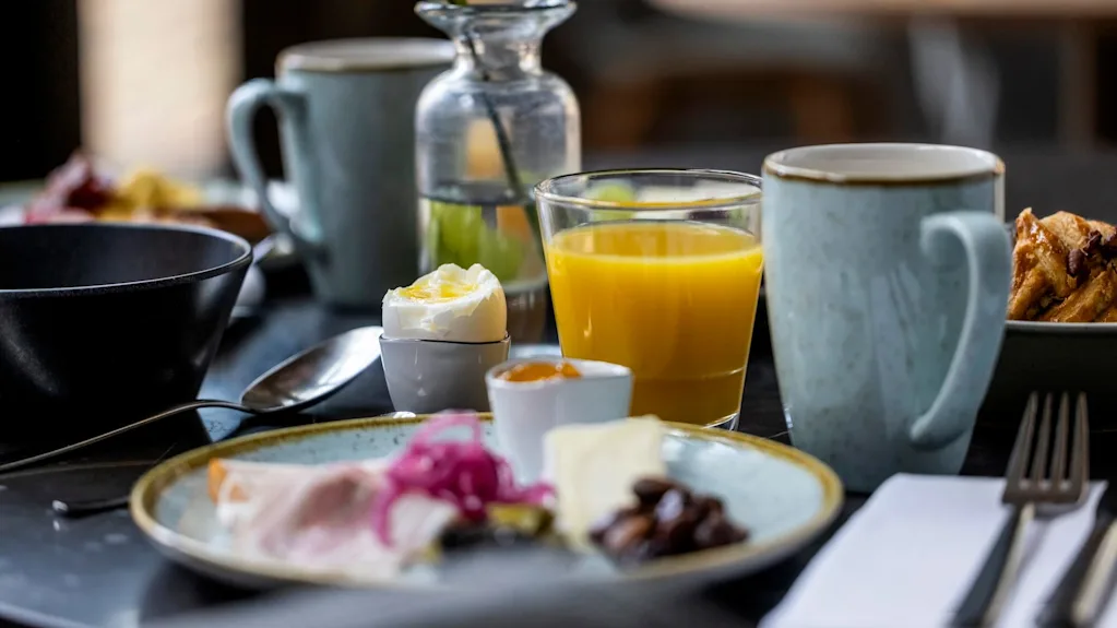 En frukostscen med en tallrik med olika maträtter, ett kokt ägg, ett glas apelsinjuice och en mugg på ett bord, i en avslappnad matmiljö.