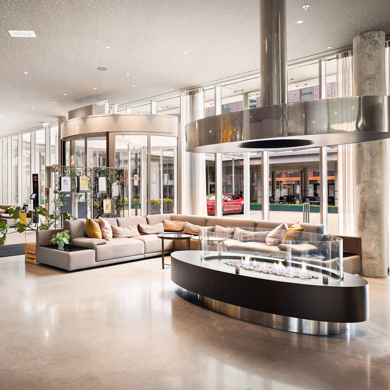 Luxurious sofa sits by a glass fireplace in a spacious, modern hotel lobby with large windows and potted plants. A sign reads, "It's all about you."
