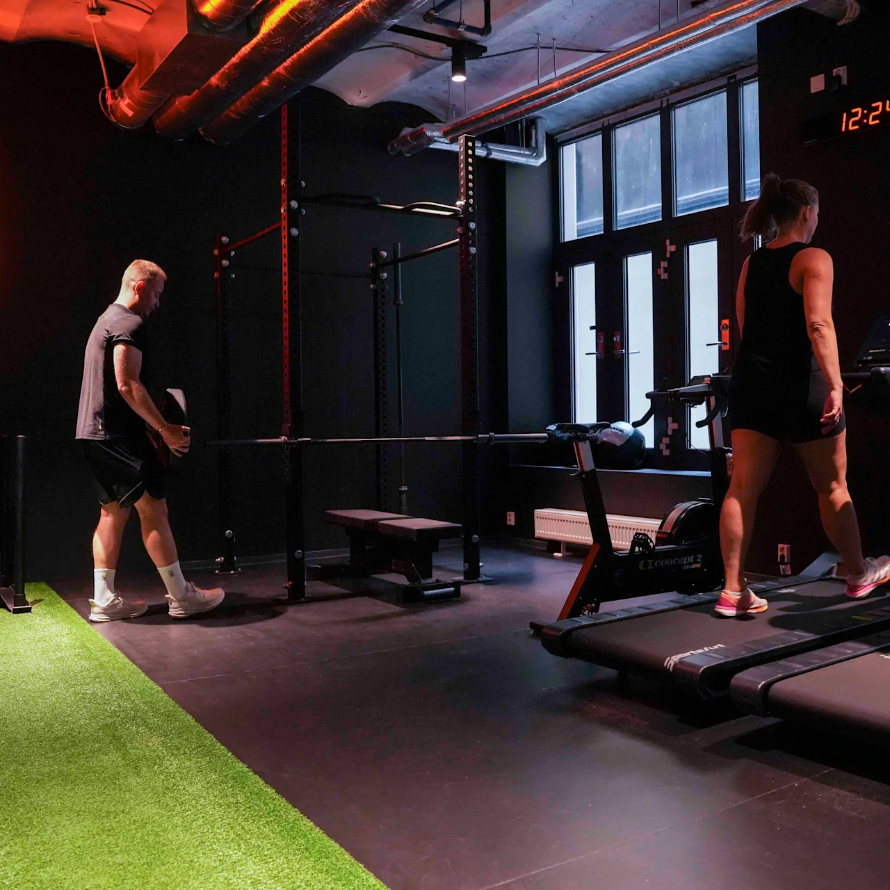 Et treningssenter med folk som trener på tredemøller og en gressstripe, med et neonskilt med teksten "ONE MORE REP" og en klokke som viser 12:24.