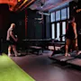 Et treningssenter med folk som trener på tredemøller og en gressstripe, med et neonskilt med teksten "ONE MORE REP" og en klokke som viser 12:24.