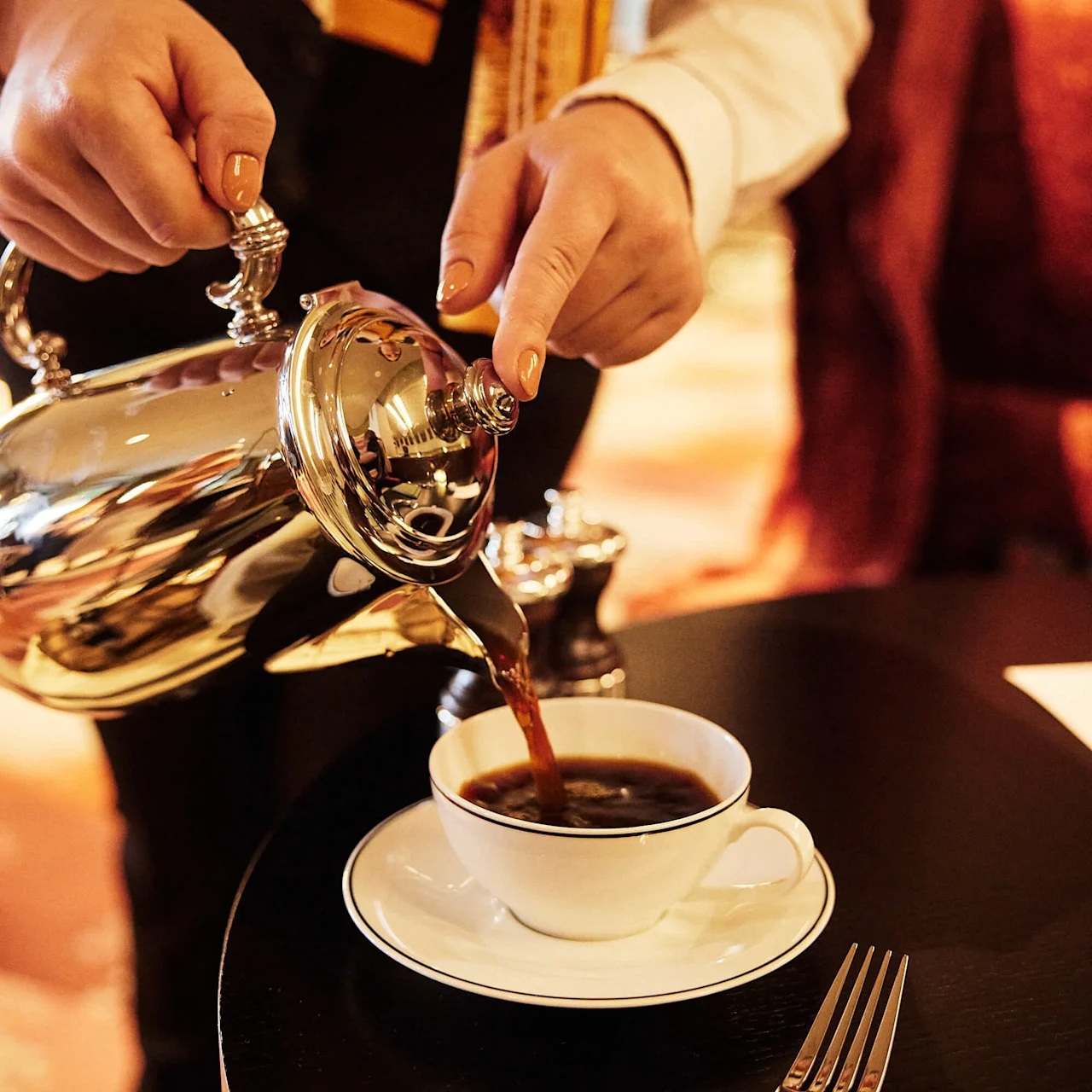 En person hælder kaffe i en kop fra en sølvkande på et bord med en mørk baggrund.