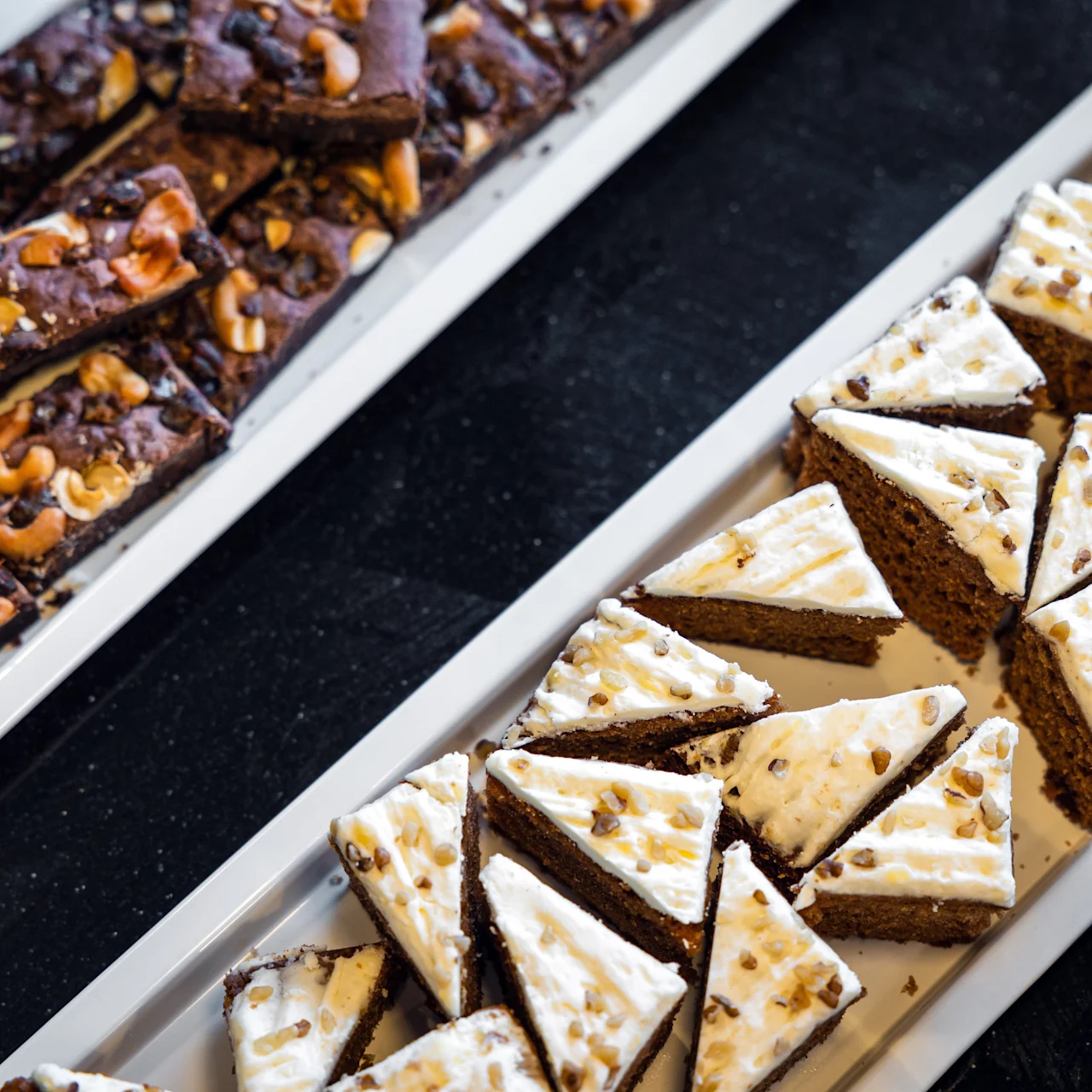 Brownies cashewpähkinöillä ja kolmionmuotoiset kuorrutetut kakut, joiden päällä on pähkinöitä, aseteltuina valkoisille tarjoittimille tummalle pinnalle.