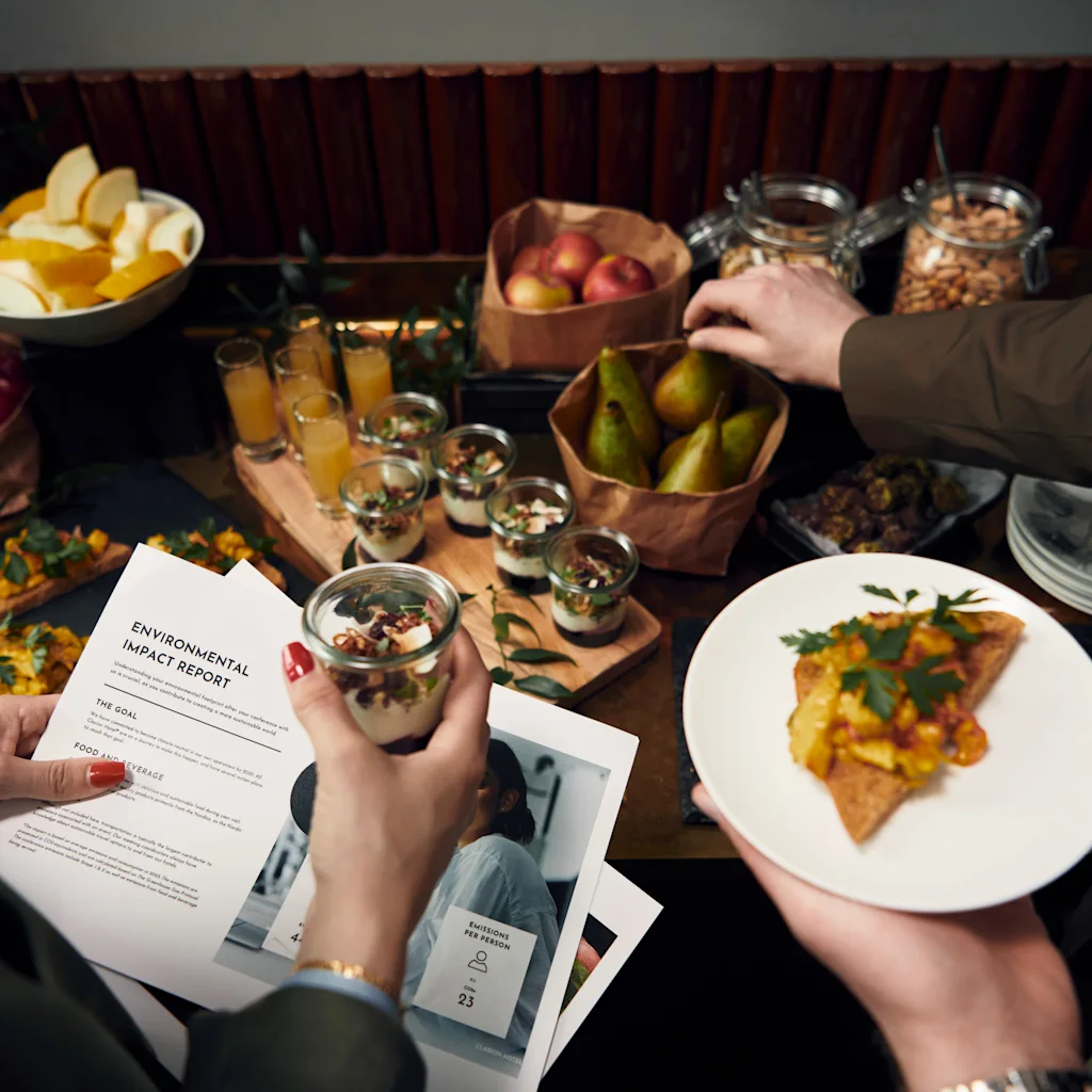 En person holder en liten dessertkopp og leser et dokument med tittelen "ENVIRONMENTAL IMPACT REPORT" ved siden av et buffetbord med frukt og appetittvekkere. En annen person velger ting fra bordet.