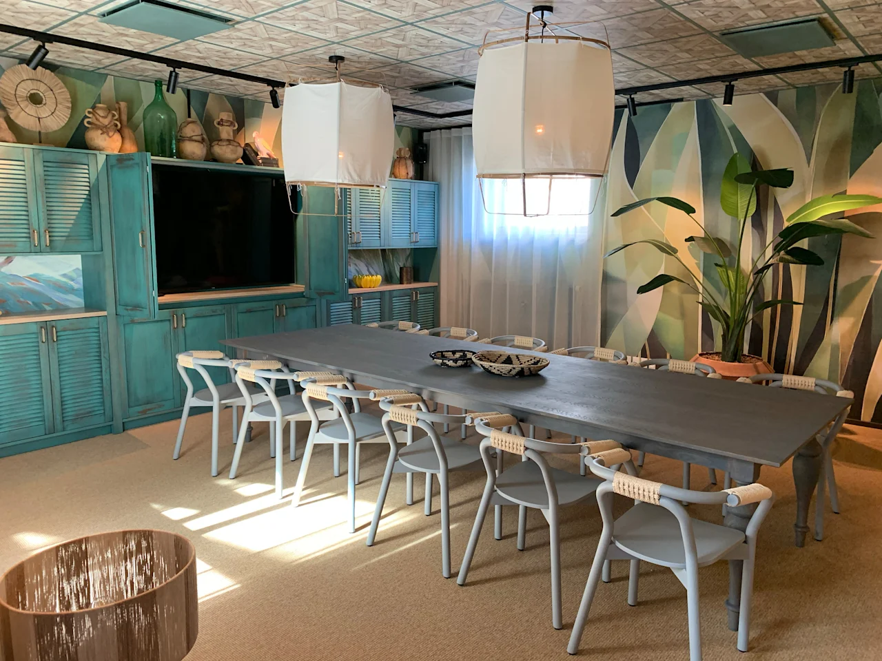 A long dining table is surrounded by white chairs, set under two large ceiling lamps. The room features turquoise cabinetry, abstract wall art, potted plants, and soft lighting.