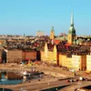 Farverige historiske bygninger omgiver en travl byplads med mennesker, der går ved en reflekterende vandvej. Spir og tårne hæver sig mod en klar blå himmel og skaber en malerisk byscene.