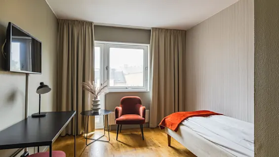 Et koselig, minimalistisk rom med en enkeltseng dekket i hvitt og oransje, en lenestol ved vinduet og et lite skrivebord med en lampe. Naturlig lys filtrerer inn gjennom gardinene og skaper en rolig atmosfære.